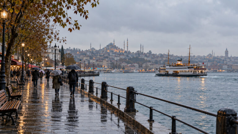 Islak sahil yolunda şemsiyeli yayalar ve denizde ilerleyen bir vapur.