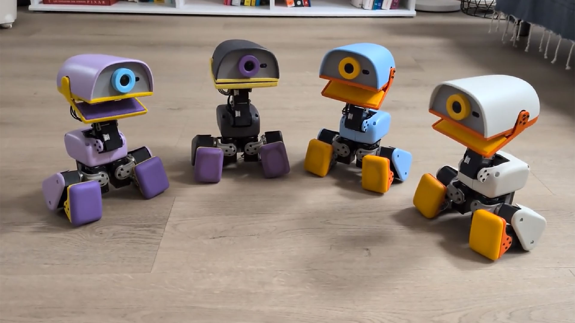 Ahşap zeminde yan yana oturan mor, koyu gri, mavi ve krem renkli dört Microduck robotu